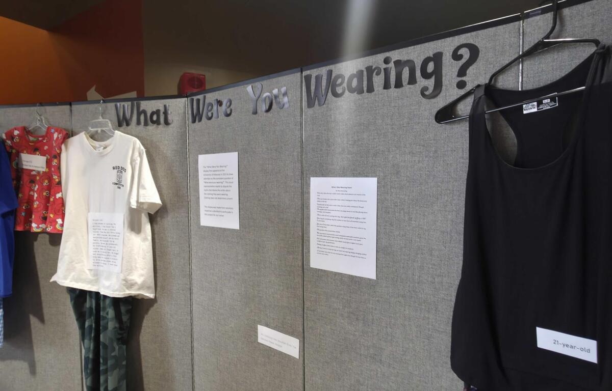Exhibit wall with clothing on hangers and a sign reading 'What Were You Wearing?' over gray partition panels, displaying shirts and a black dress.