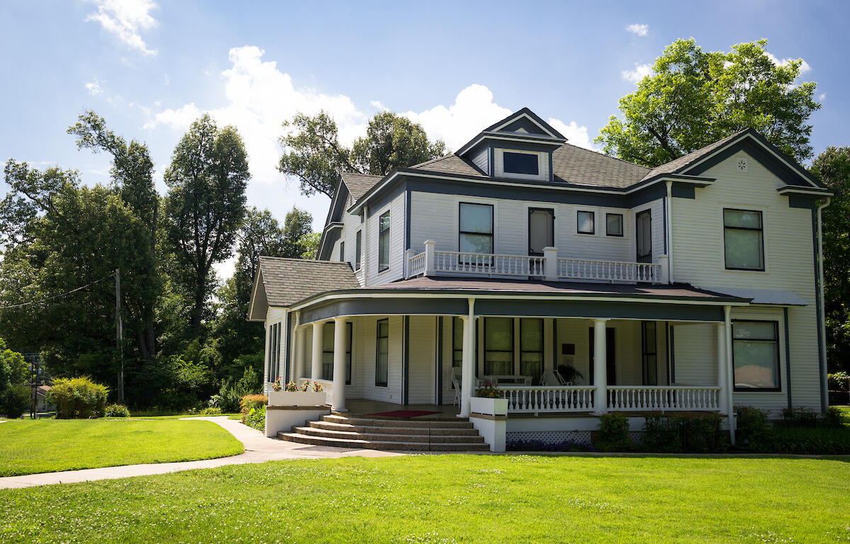 A large two-story white house with a wraparound porch, columns, and a front staircase, set on a green lawn with trees against a blue sky.