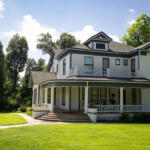 A large two-story white house with a wraparound porch, columns, and a front staircase, set on a green lawn with trees against a blue sky.