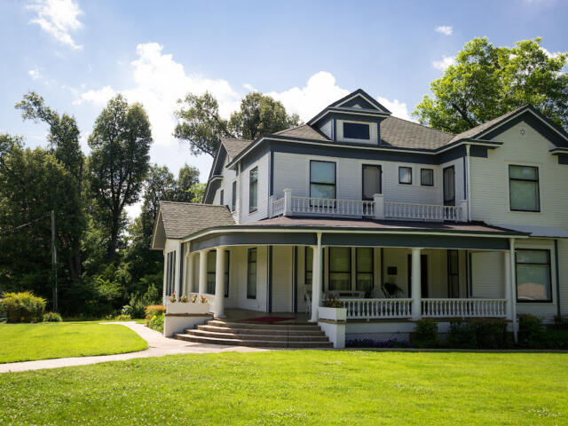 A large two-story white house with a wraparound porch, columns, and a front staircase, set on a green lawn with trees against a blue sky.