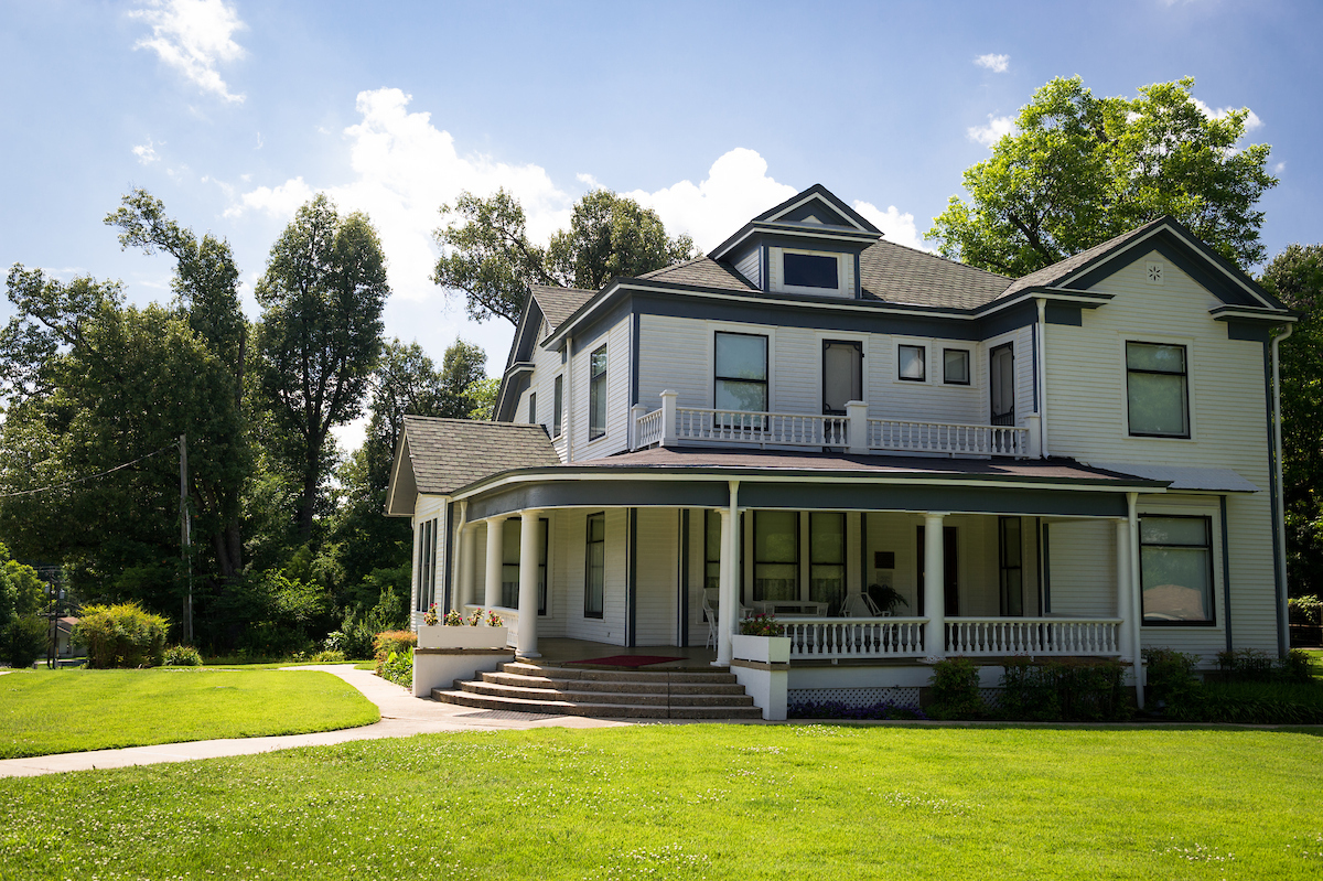 A large two-story white house with a wraparound porch, columns, and a front staircase, set on a green lawn with trees against a blue sky.