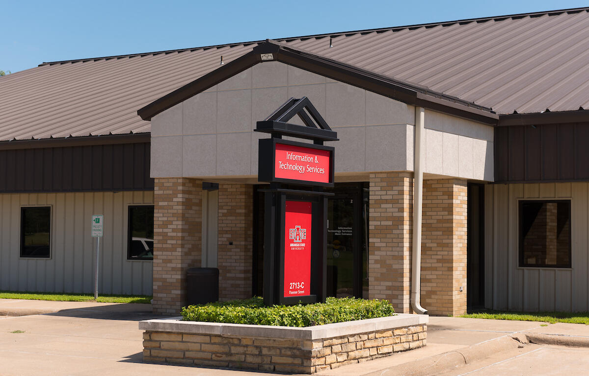 Entrance to Information & Technology Services at a campus building with a red sign in a black frame, beige brick columns and a raised garden bed in front.