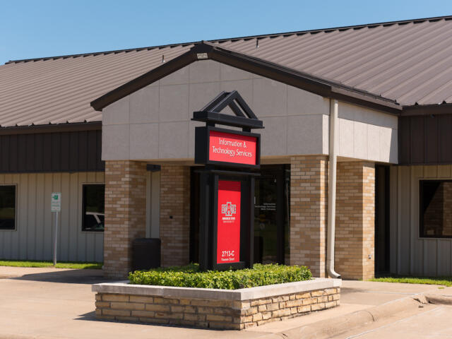 Entrance to Information & Technology Services at a campus building with a red sign in a black frame, beige brick columns and a raised garden bed in front.