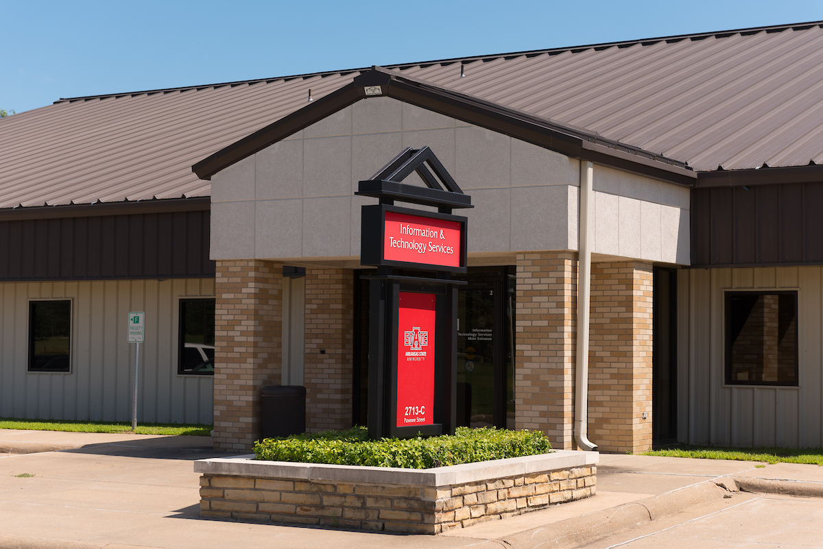 Entrance to Information & Technology Services at a campus building with a red sign in a black frame, beige brick columns and a raised garden bed in front.