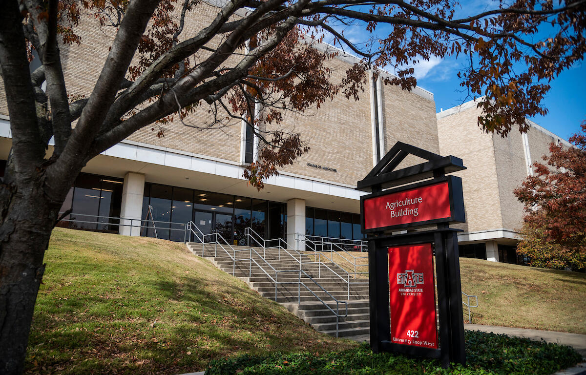 Exterior view of the Agriculture Building at Arkansas State University, a red sign in the foreground, brick facade, stairs, and blue sky framed by trees.