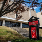 Exterior view of the Agriculture Building at Arkansas State University, a red sign in the foreground, brick facade, stairs, and blue sky framed by trees.