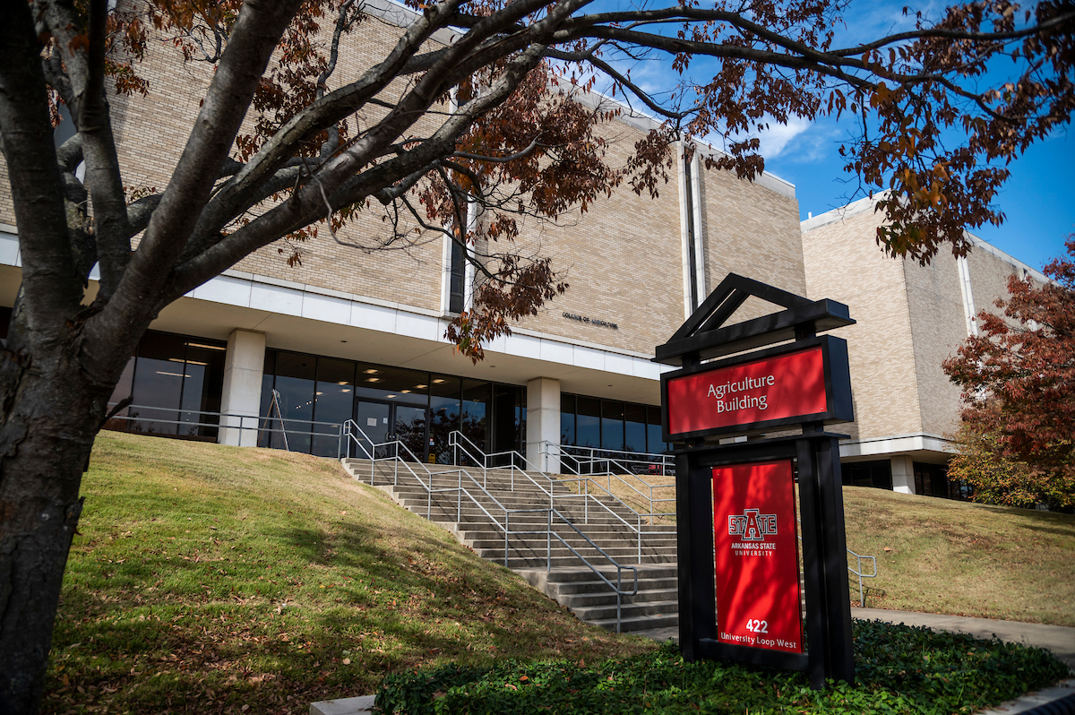 Exterior view of the Agriculture Building at Arkansas State University, a red sign in the foreground, brick facade, stairs, and blue sky framed by trees.