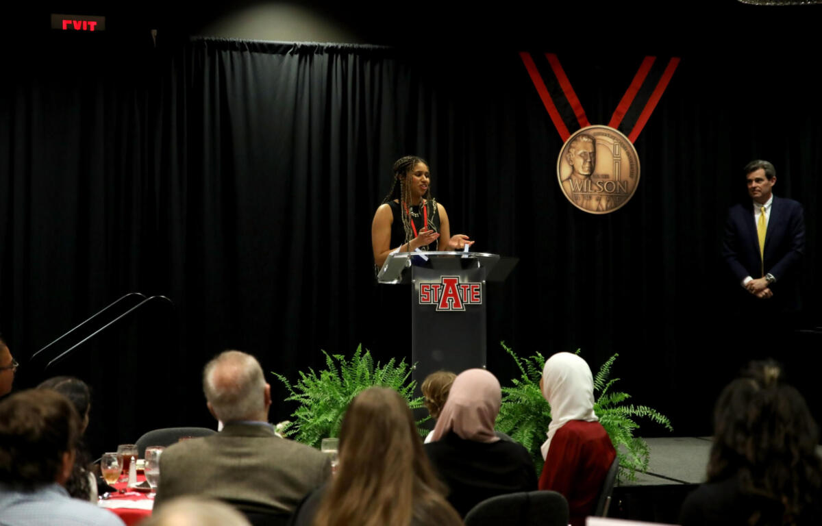 Woman speaks at a podium on a dark stage, STATE logo on the podium, audience watching, with a man in a suit standing to the right.