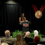 Woman speaks at a podium on a dark stage, STATE logo on the podium, audience watching, with a man in a suit standing to the right.