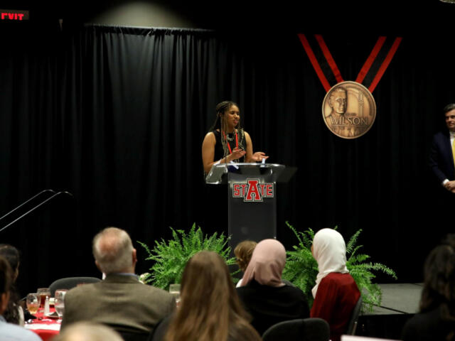 Woman speaks at a podium on a dark stage, STATE logo on the podium, audience watching, with a man in a suit standing to the right.
