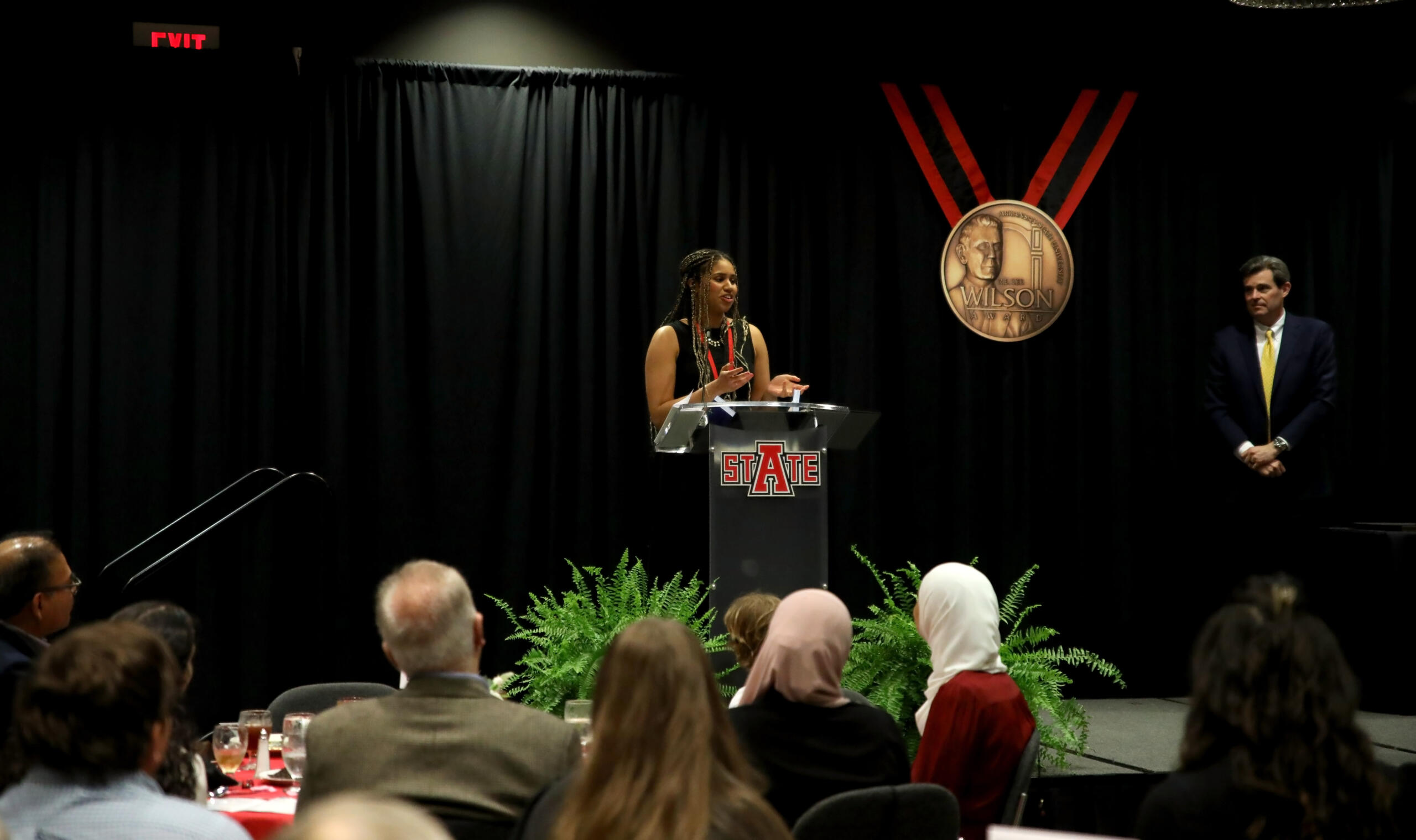 Woman speaks at a podium on a dark stage, STATE logo on the podium, audience watching, with a man in a suit standing to the right.