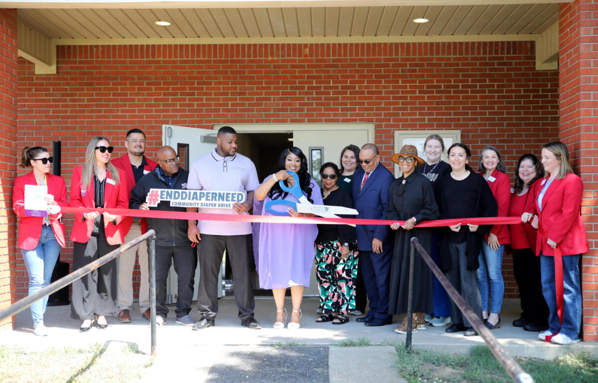 Group of diverse adults at a ribbon-cutting event outside a brick building, holding a red ribbon and a sign about diaper drives.
