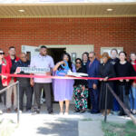 Group of diverse adults at a ribbon-cutting event outside a brick building, holding a red ribbon and a sign about diaper drives.