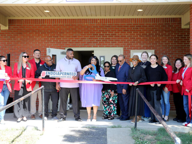 Group of diverse adults at a ribbon-cutting event outside a brick building, holding a red ribbon and a sign about diaper drives.