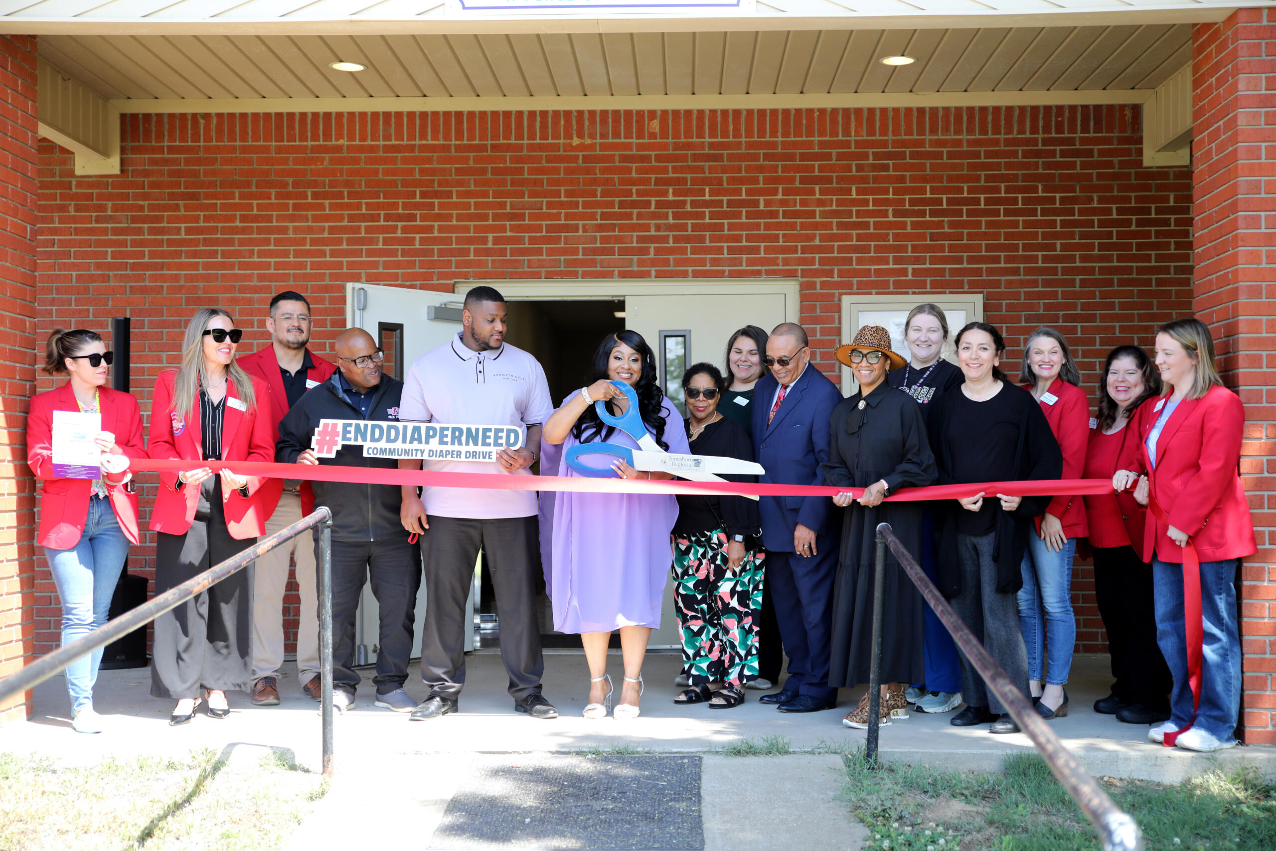 Group of diverse adults at a ribbon-cutting event outside a brick building, holding a red ribbon and a sign about diaper drives.