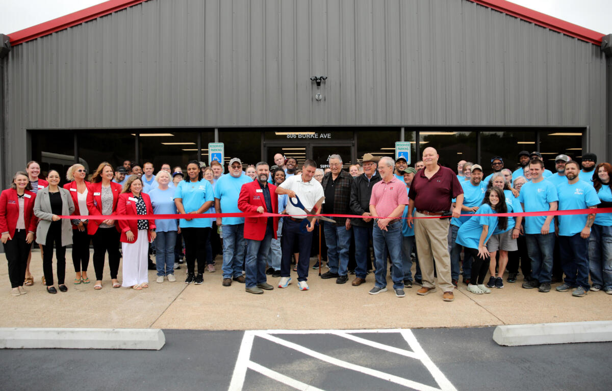 Large group of people gathered for a ribbon-cutting in front of a gray metal building with a red roof.",