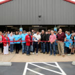 Large group of people gathered for a ribbon-cutting in front of a gray metal building with a red roof.",