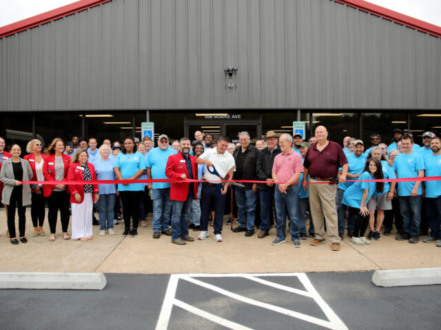 Large group of people gathered for a ribbon-cutting in front of a gray metal building with a red roof.",