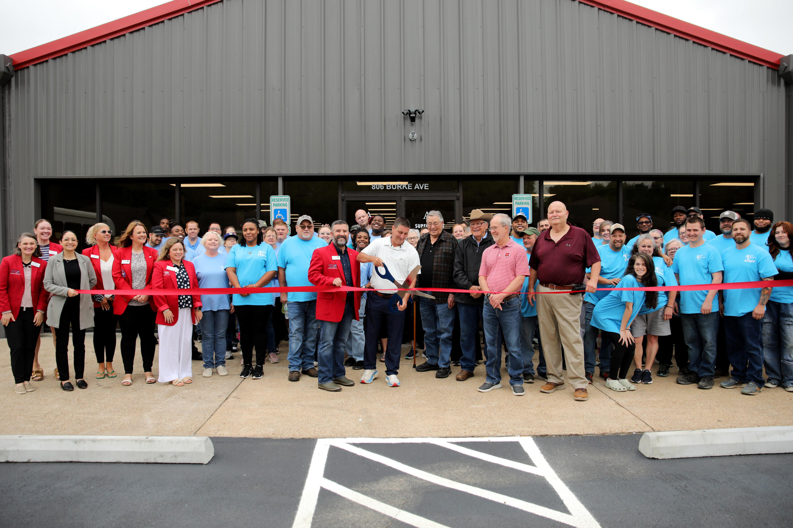 Large group of people gathered for a ribbon-cutting in front of a gray metal building with a red roof.",