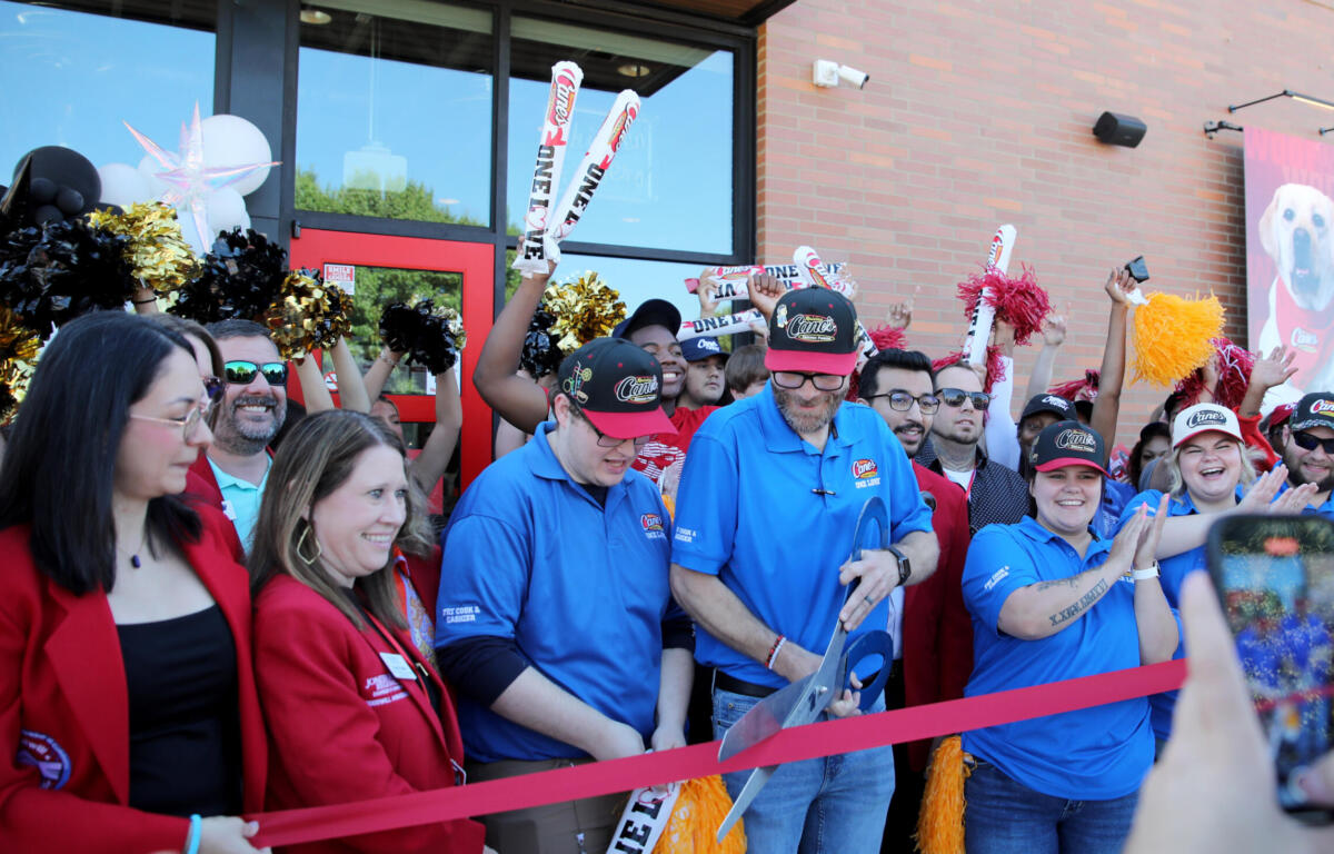 Crowd of staff and guests at a Cane's restaurant opening, cheering behind a red ribbon outside a brick building with cane's branding.