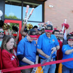 Crowd of staff and guests at a Cane's restaurant opening, cheering behind a red ribbon outside a brick building with cane's branding.