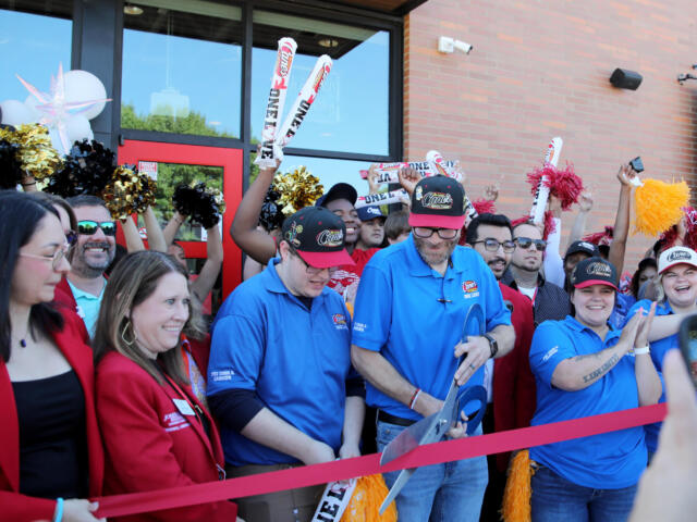 Crowd of staff and guests at a Cane's restaurant opening, cheering behind a red ribbon outside a brick building with cane's branding.