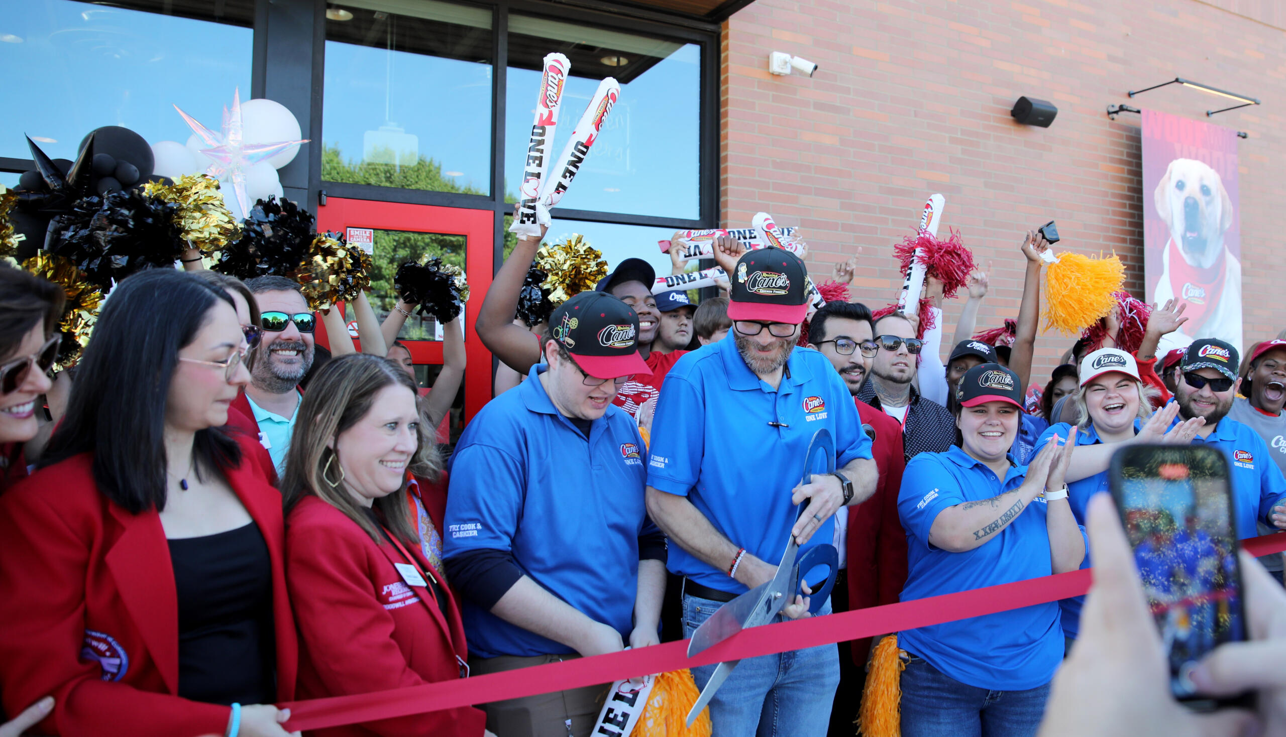 Crowd of staff and guests at a Cane's restaurant opening, cheering behind a red ribbon outside a brick building with cane's branding.