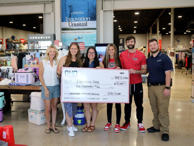Six smiling people in a sporting goods store pose with a giant $10,000 check.