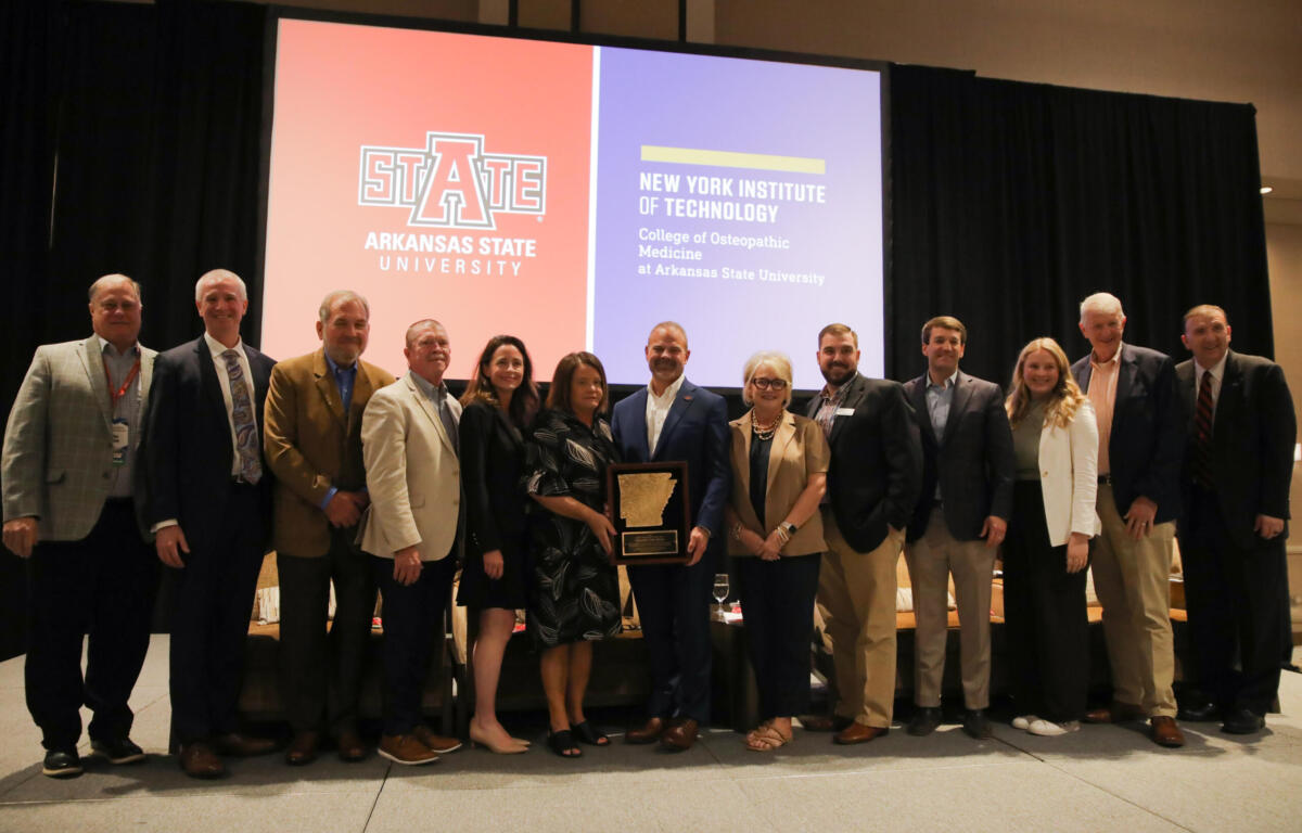 Group of professionals posing on a stage with an award plaque, in front of orange and purple banners for Arkansas State University and New York Institute of Technology.
