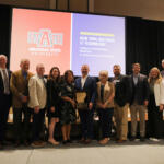 Group of professionals posing on a stage with an award plaque, in front of orange and purple banners for Arkansas State University and New York Institute of Technology.