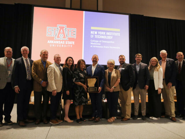 Group of professionals posing on a stage with an award plaque, in front of orange and purple banners for Arkansas State University and New York Institute of Technology.