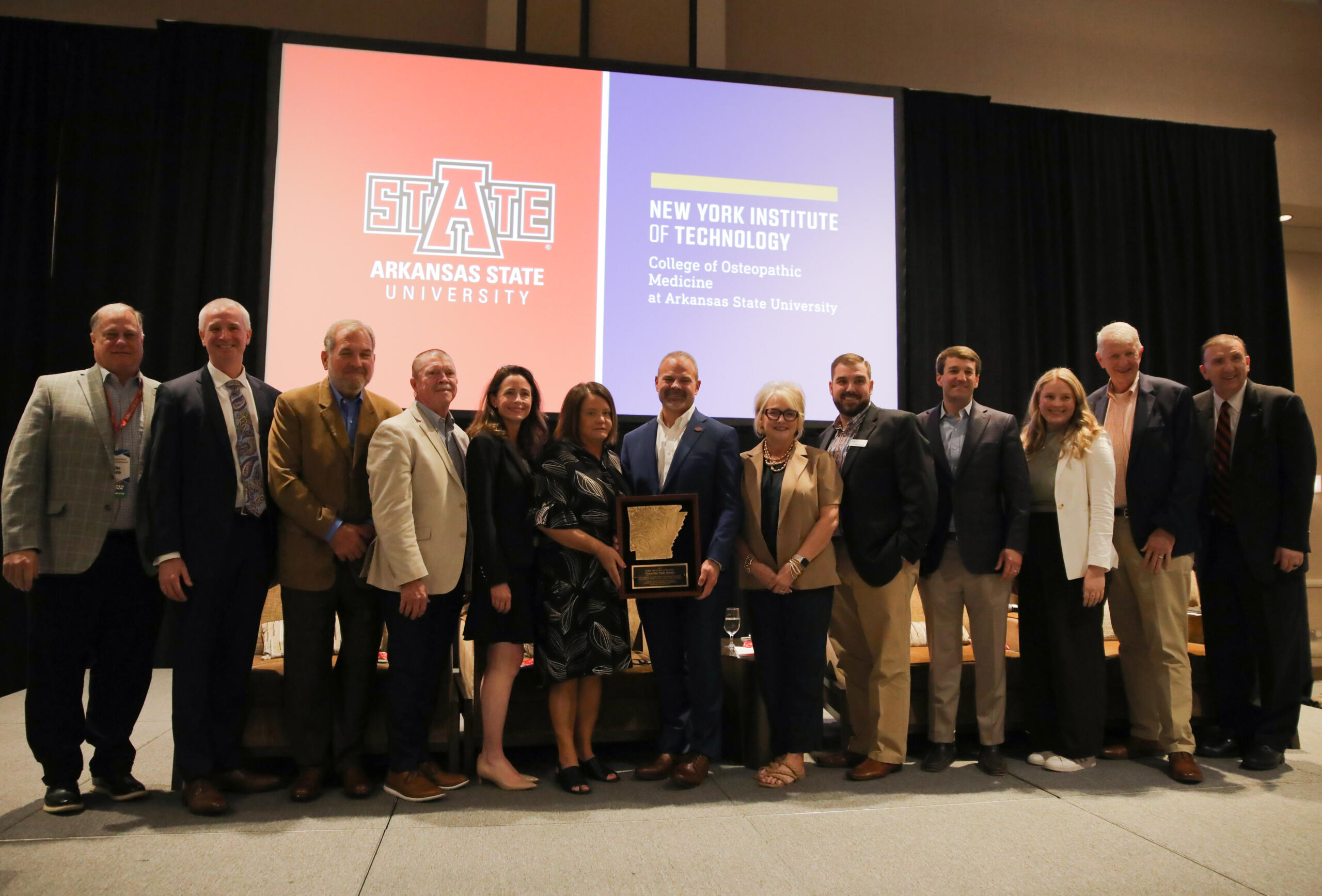 Group of professionals posing on a stage with an award plaque, in front of orange and purple banners for Arkansas State University and New York Institute of Technology.
