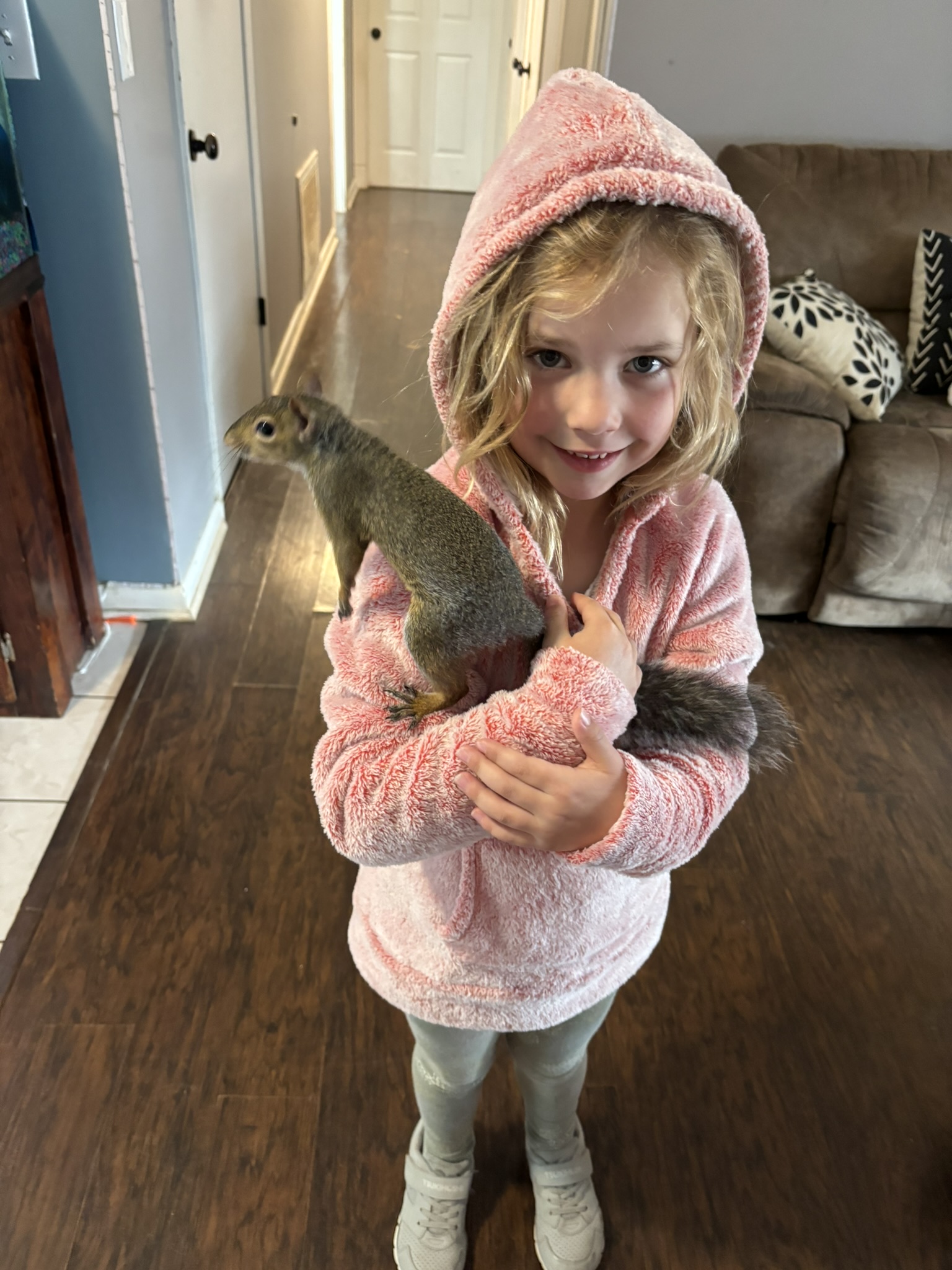Little girl in a pink bathrobe holding a gray squirrel indoors, smiling at the camera in a living room.