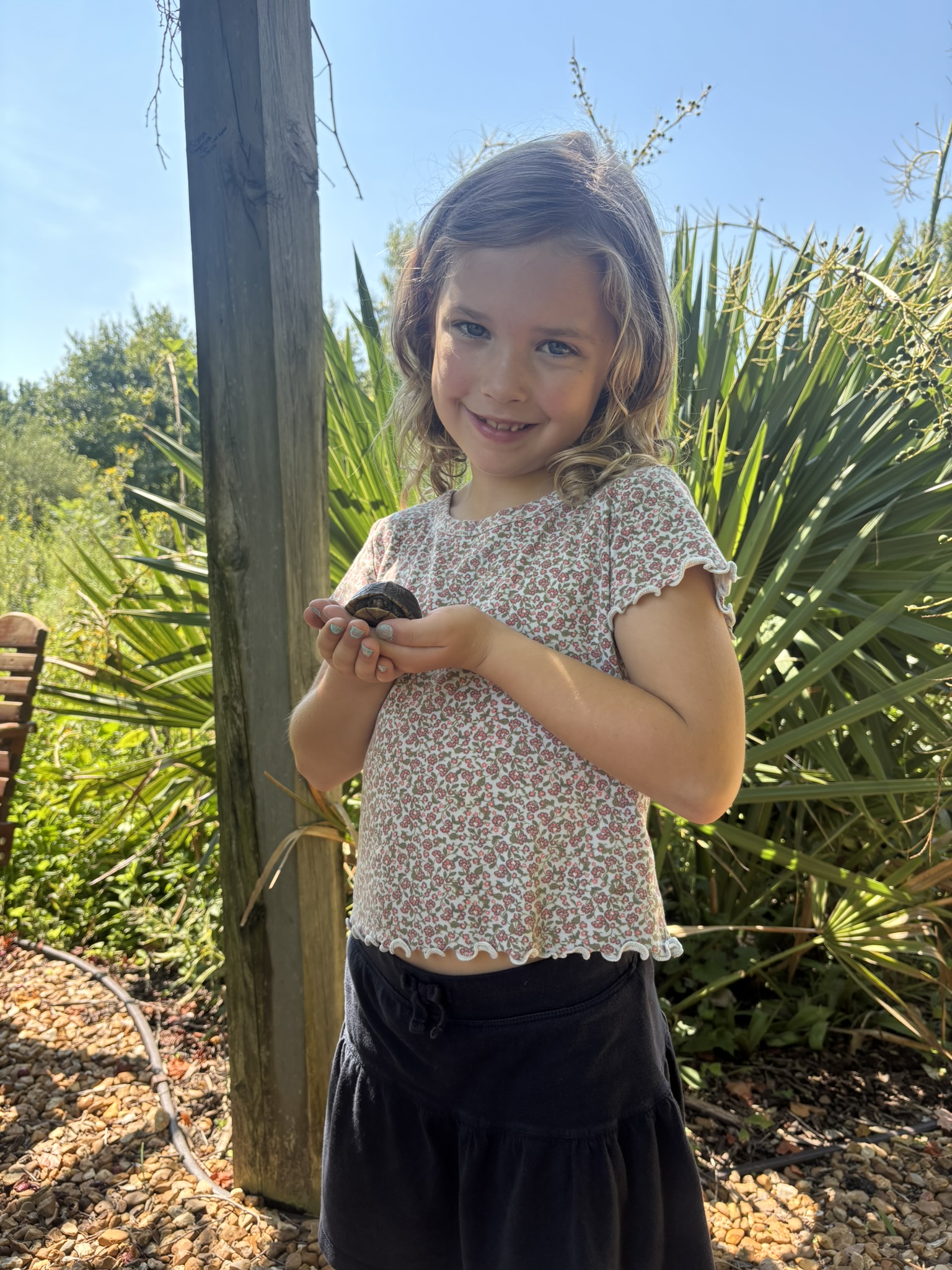 Smiling young girl holding a seashell outdoors, with green plants and a wooden post behind her.