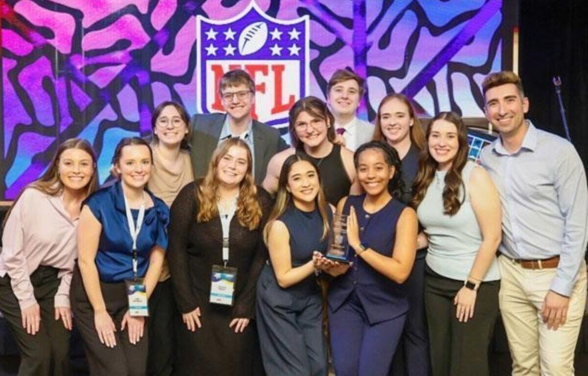 Group of 12 people posing on a stage with a trophy, smiling after an award ceremony, NFL logo backdrop in colorful art.
