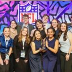 Group of 12 people posing on a stage with a trophy, smiling after an award ceremony, NFL logo backdrop in colorful art.