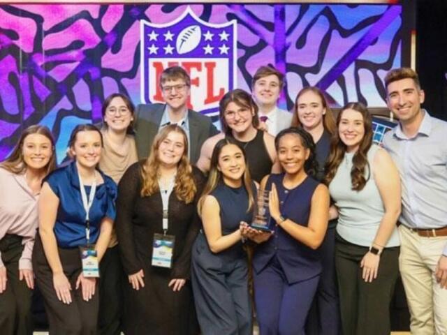 Group of 12 people posing on a stage with a trophy, smiling after an award ceremony, NFL logo backdrop in colorful art.