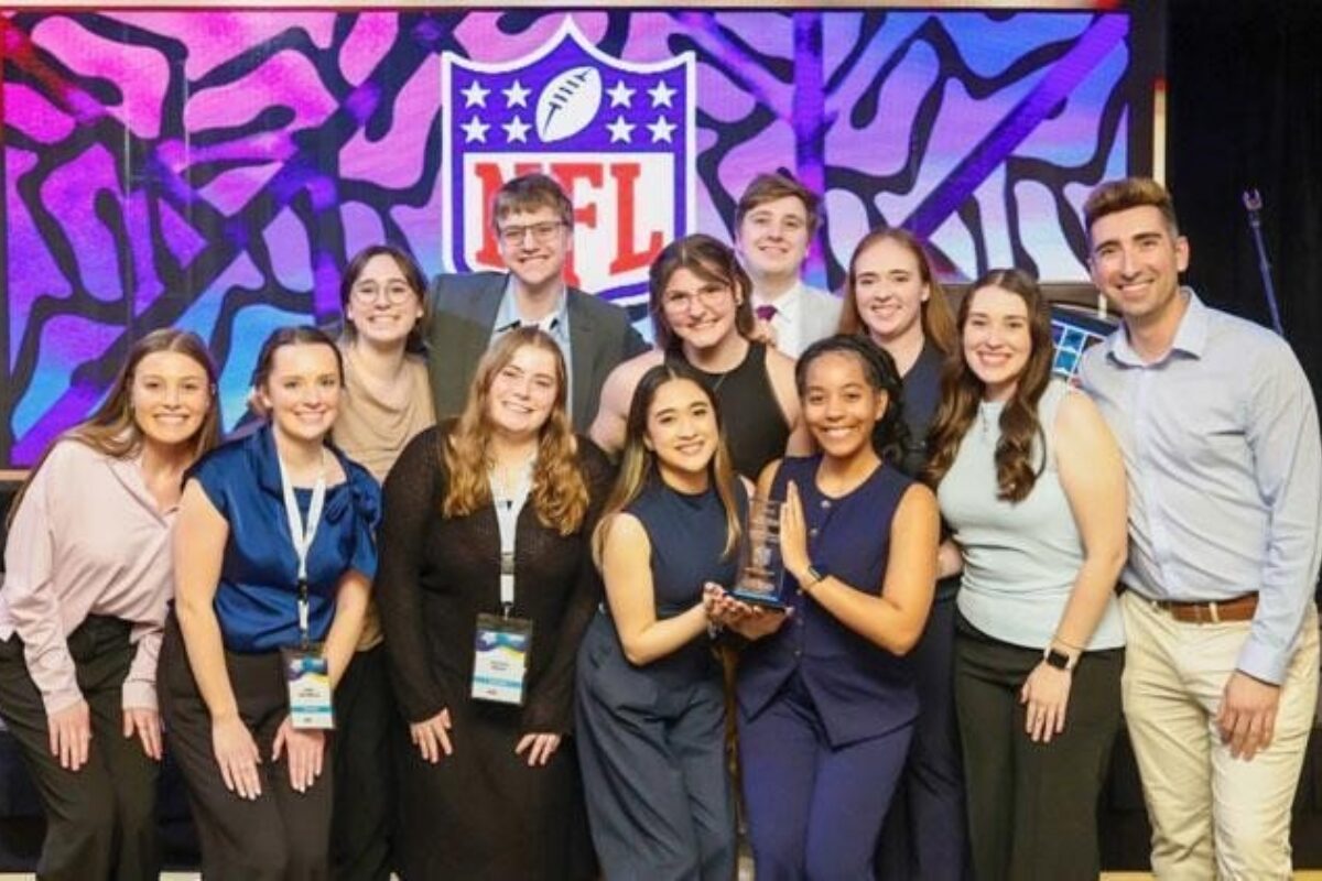 Group of 12 people posing on a stage with a trophy, smiling after an award ceremony, NFL logo backdrop in colorful art.