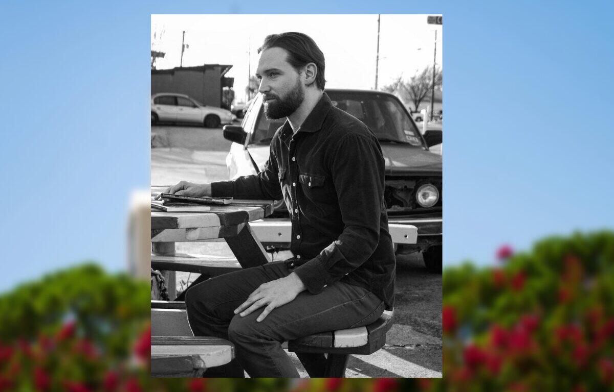 Bearded man in a dark shirt sits at a portable drafting table outdoors, focused on his work near a car in the background.