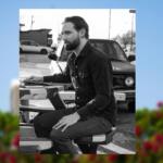 Bearded man in a dark shirt sits at a portable drafting table outdoors, focused on his work near a car in the background.