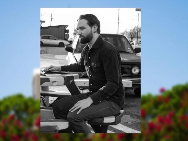 Bearded man in a dark shirt sits at a portable drafting table outdoors, focused on his work near a car in the background.
