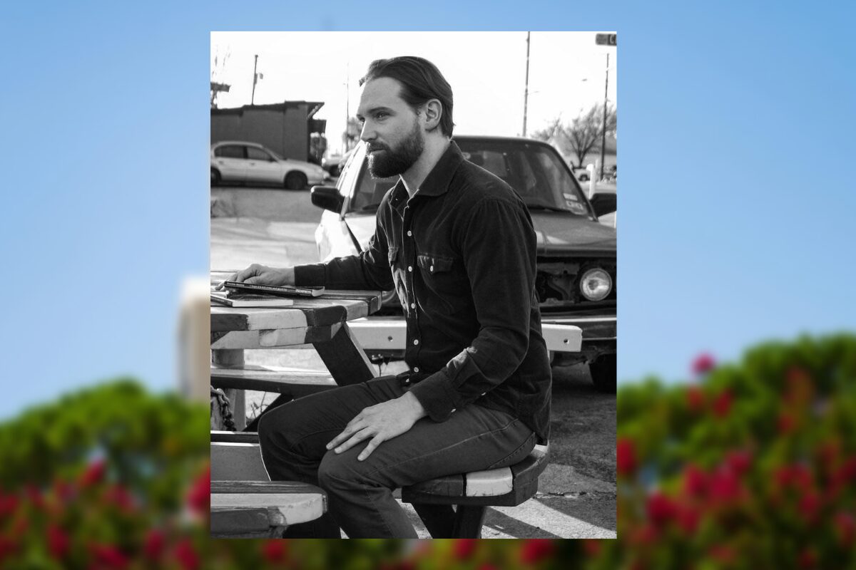 Bearded man in a dark shirt sits at a portable drafting table outdoors, focused on his work near a car in the background.
