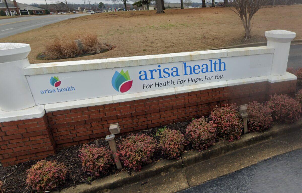 Arisa Health sign on a white wall with blue logo and slogan, set on a brick planter by a road.