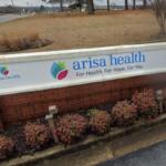 Arisa Health sign on a white wall with blue logo and slogan, set on a brick planter by a road.