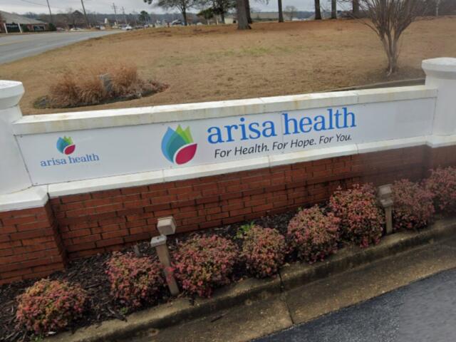 Arisa Health sign on a white wall with blue logo and slogan, set on a brick planter by a road.