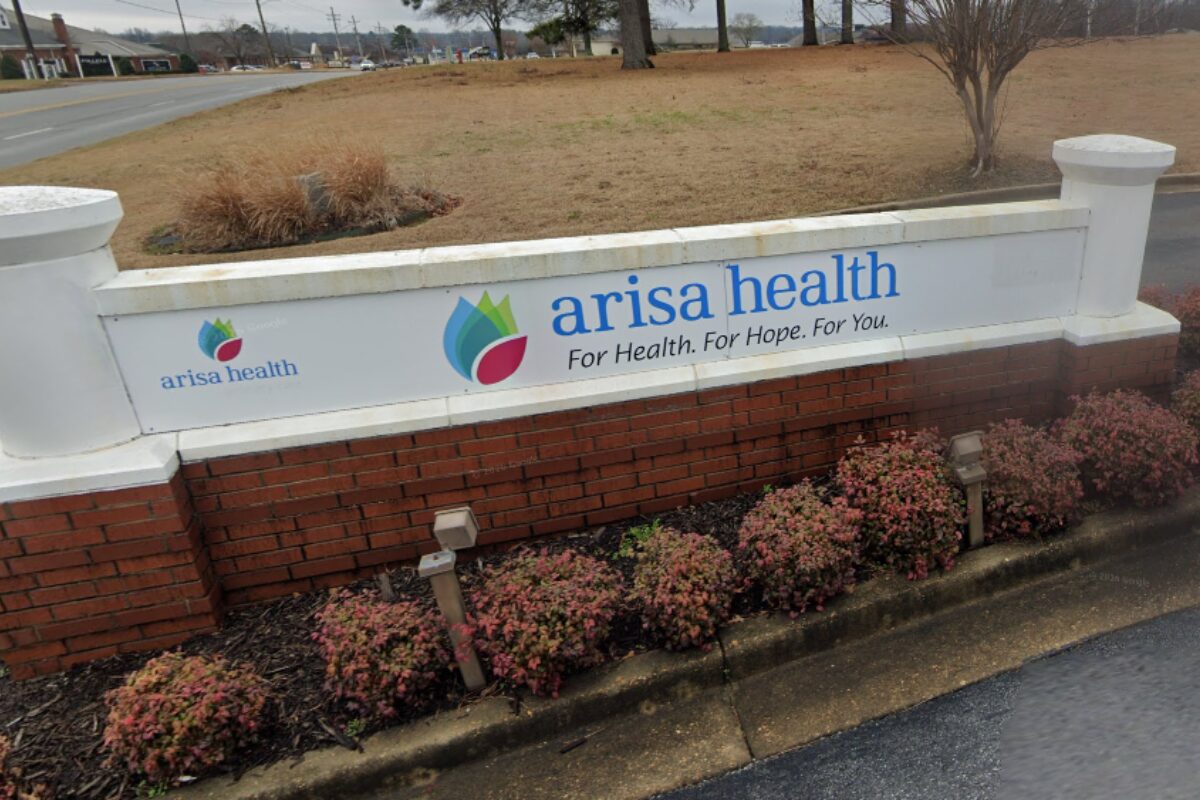 Arisa Health sign on a white wall with blue logo and slogan, set on a brick planter by a road.