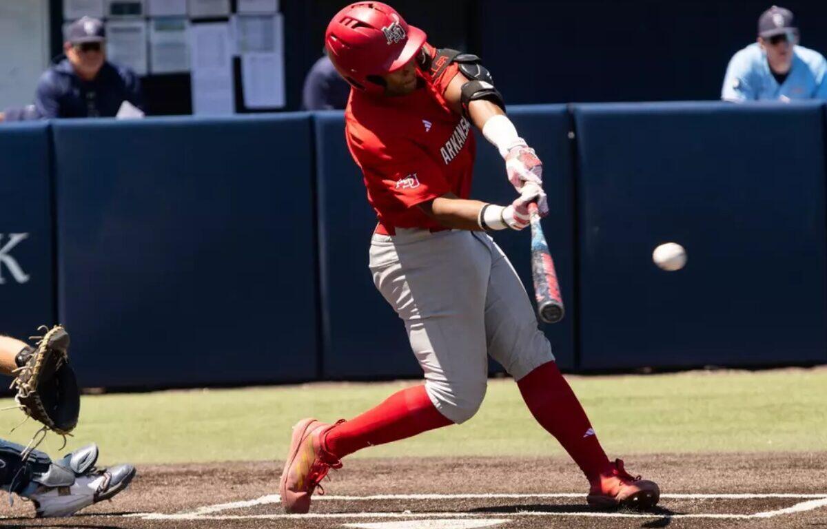 Baseball batter in red uniform mid-swing at a pitched ball near home plate, with catcher crouched behind him.