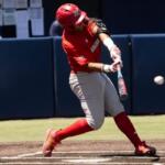 Baseball batter in red uniform mid-swing at a pitched ball near home plate, with catcher crouched behind him.