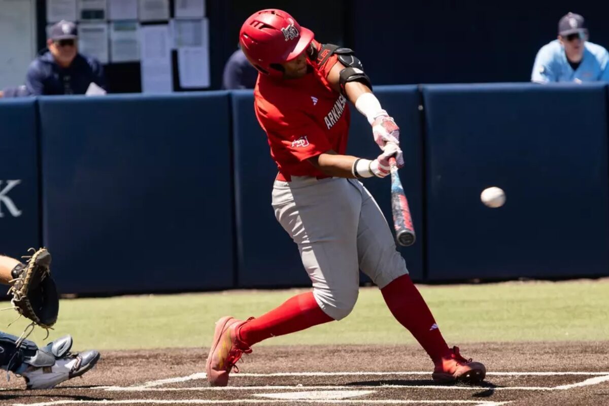 Baseball batter in red uniform mid-swing at a pitched ball near home plate, with catcher crouched behind him.