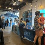 Indoor community event with multiple info booths, string lights, and people browsing tables along the room.